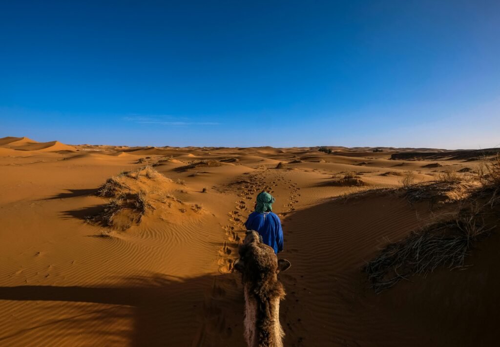 Person riding a camel through the dunes of Ksar Tanamouste, Morocco under a clear blue sky.